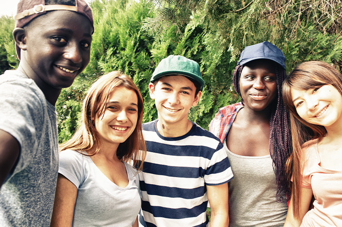 Ein Bild, das fröhliche junge Menschen unterschiedlicher ethnischer Herkunft zeigt, die ein Selfie vor einem Nadelbaum machen.