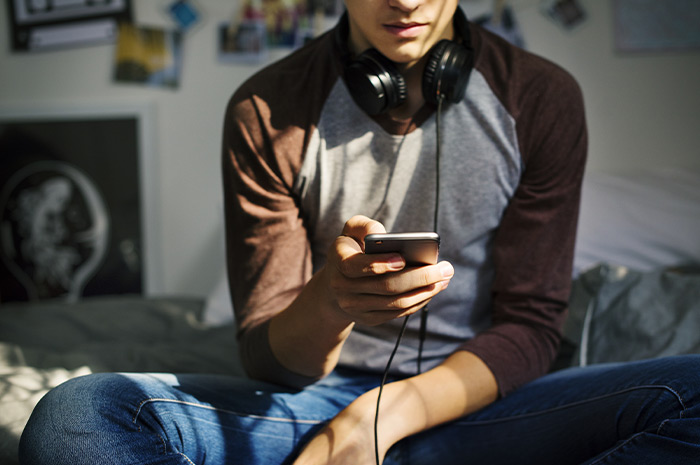 Ein Bild, das einen Jungen zeigt, der mit Kopfhörern und Smartphone auf seinem Bett sitzt.